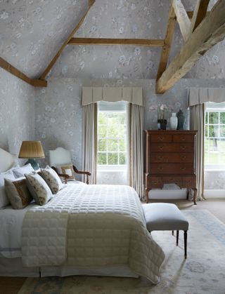 grey and white bedroom with grey floral wallpaper, wooden beams, vintage tall ball, pelmet drapes, armchair, ottoman