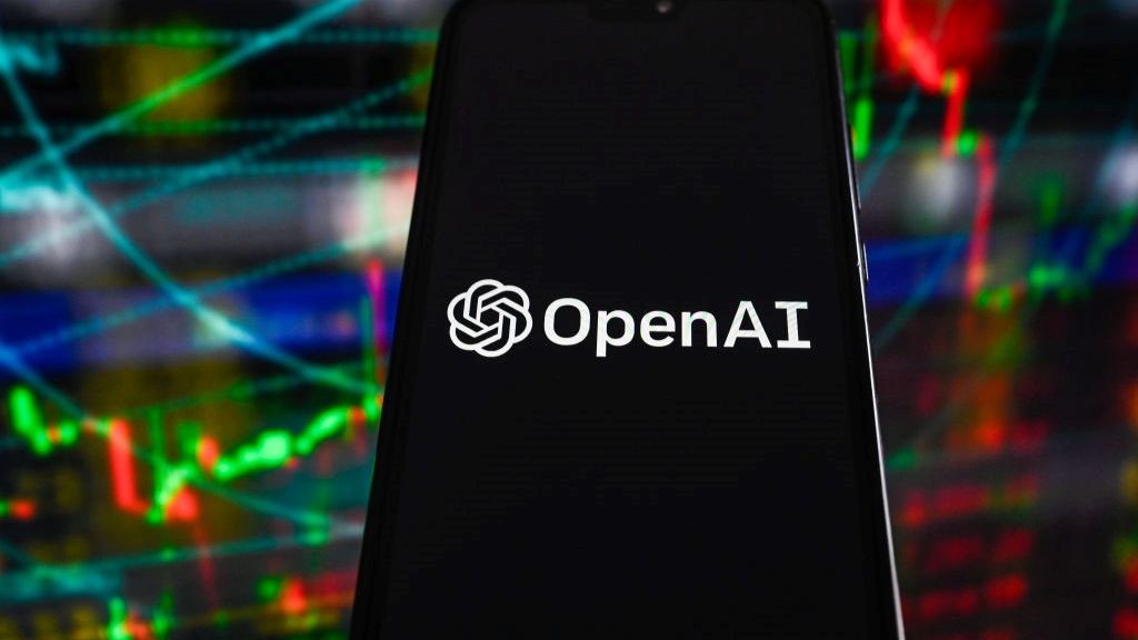 A smartphone with the words OpenAI displayed on screen, being held in front of a background showing financial charts