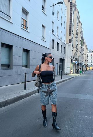 a photo showing what to wear to a rap concert with a woman wearing sporty sunglasses with a black tube top, long denim shorts, black cowboy boots, and a Balenciaga bag