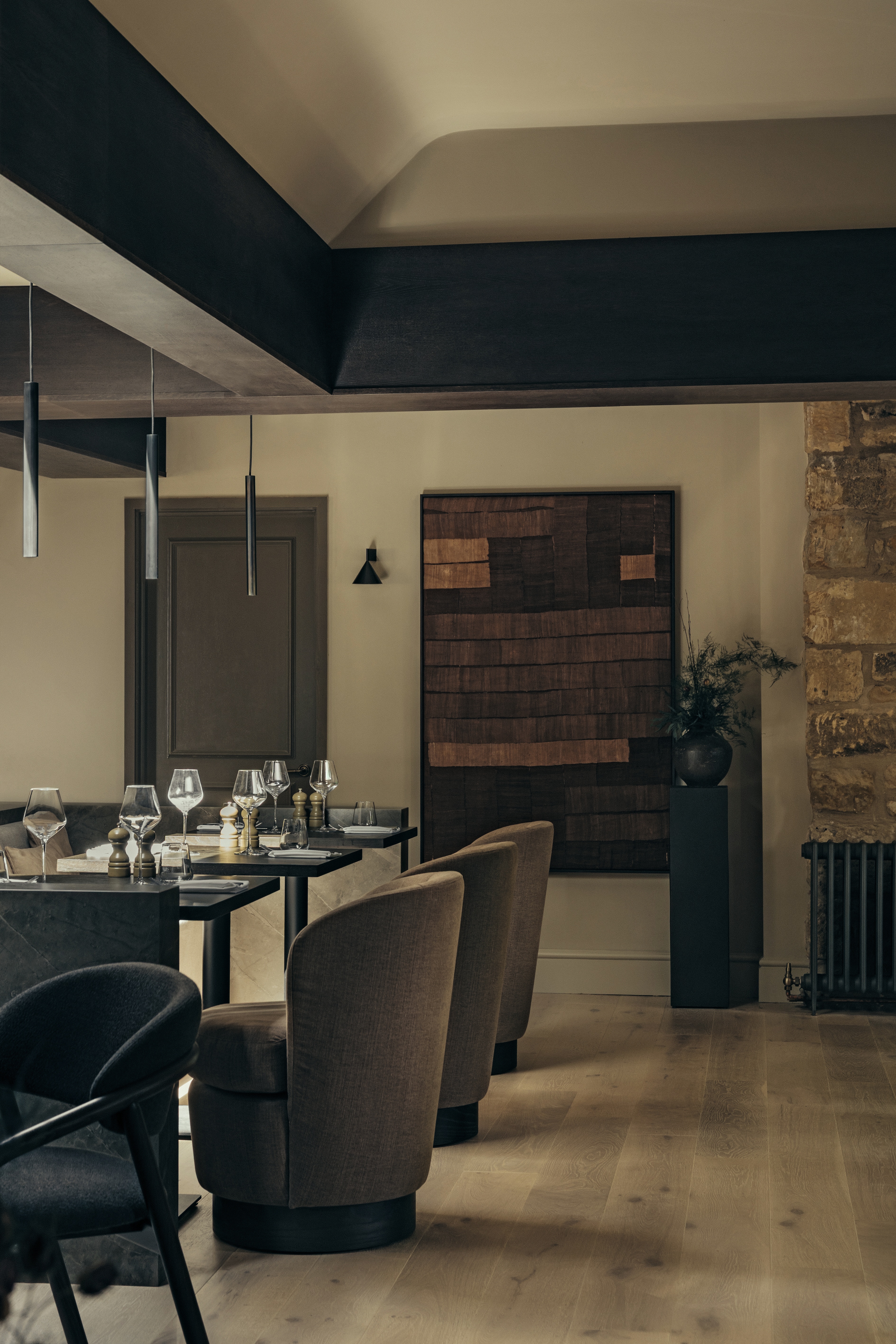 An earthy-tinted, wood-clad hotel bar boasts upholstered, brown velvet armchairs, essential black earthenware ceramics, exposed stone walls, and wooden floors.