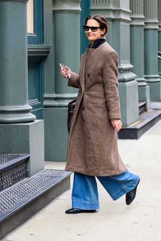 A photo of Katie Holmes styling a brown herringbone coat with wide-leg jeans, black ballet flats, and belted brown bucket bag.