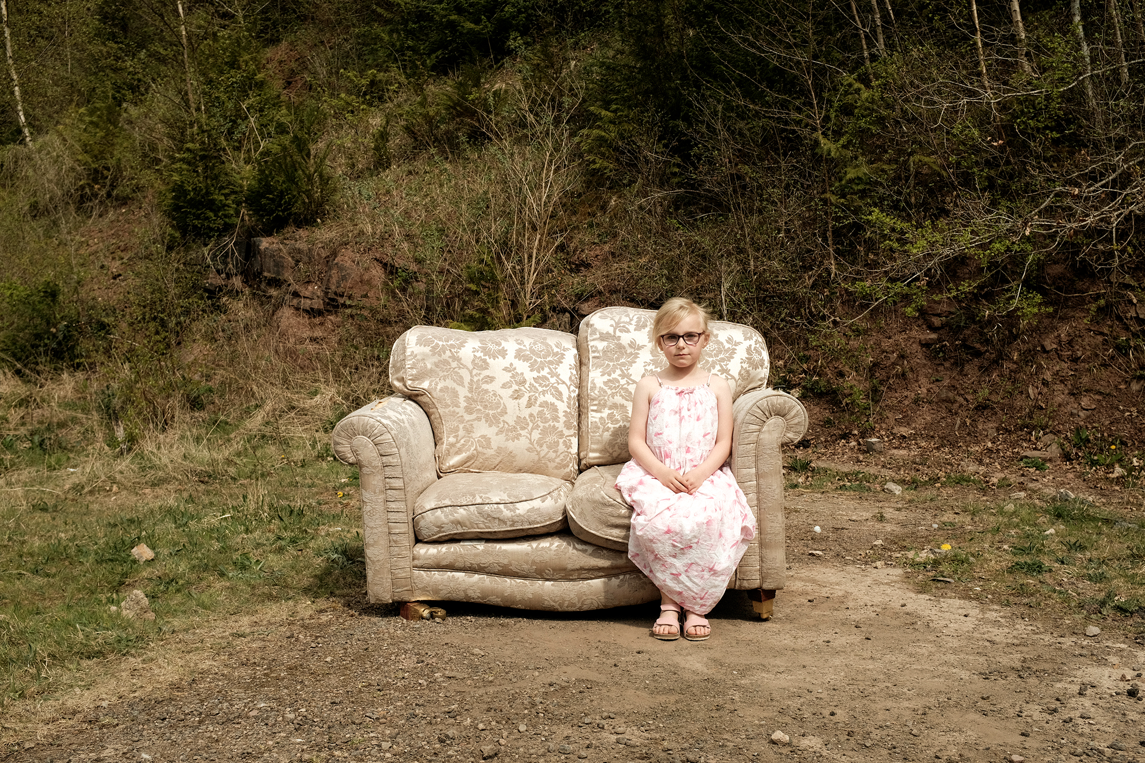 A child in a floral dress sits on a worn beige sofa placed outdoors, surrounded by greenery and dirt
