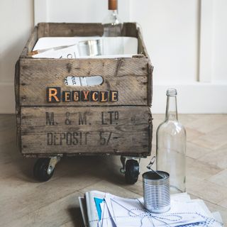 Apple crate with wheels and recycling written on the side of it, filled with paper and a glass bottle next to it