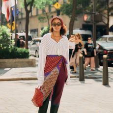 Naomi wears white button down, black trousers, red bandana, red braided handbag, flat sandals, and a scarf around her waist. 