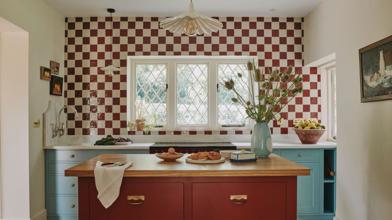 a kitchen with a checkerboard tile backsplash, lower blue painted cabinets, an island with a wooden top surface and red painted drawers, a vase of flowers on the island, and a pendant light hanging from the ceiling