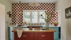a kitchen with a checkerboard tile backsplash, lower blue painted cabinets, an island with a wooden top surface and red painted drawers, a vase of flowers on the island, and a pendant light hanging from the ceiling