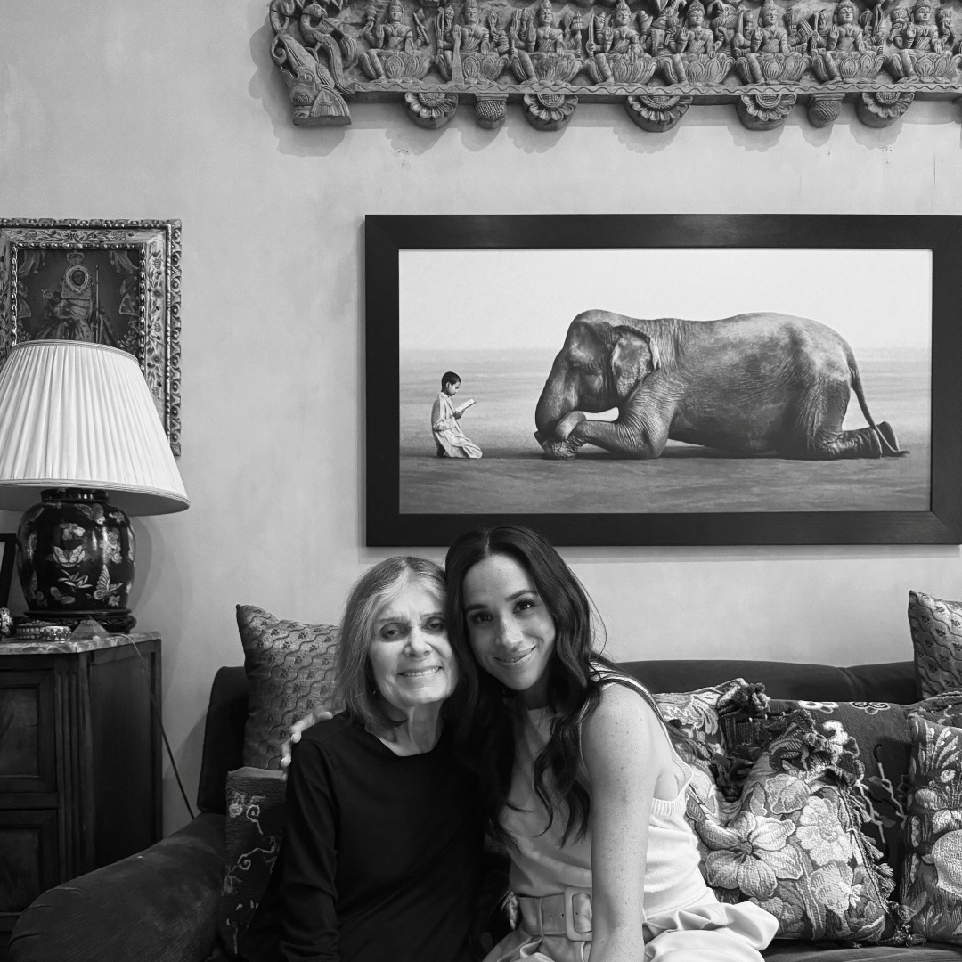 A black and white photo of Meghan Markle sitting on a sofa with Gloria Steinem in front of an elephant painting