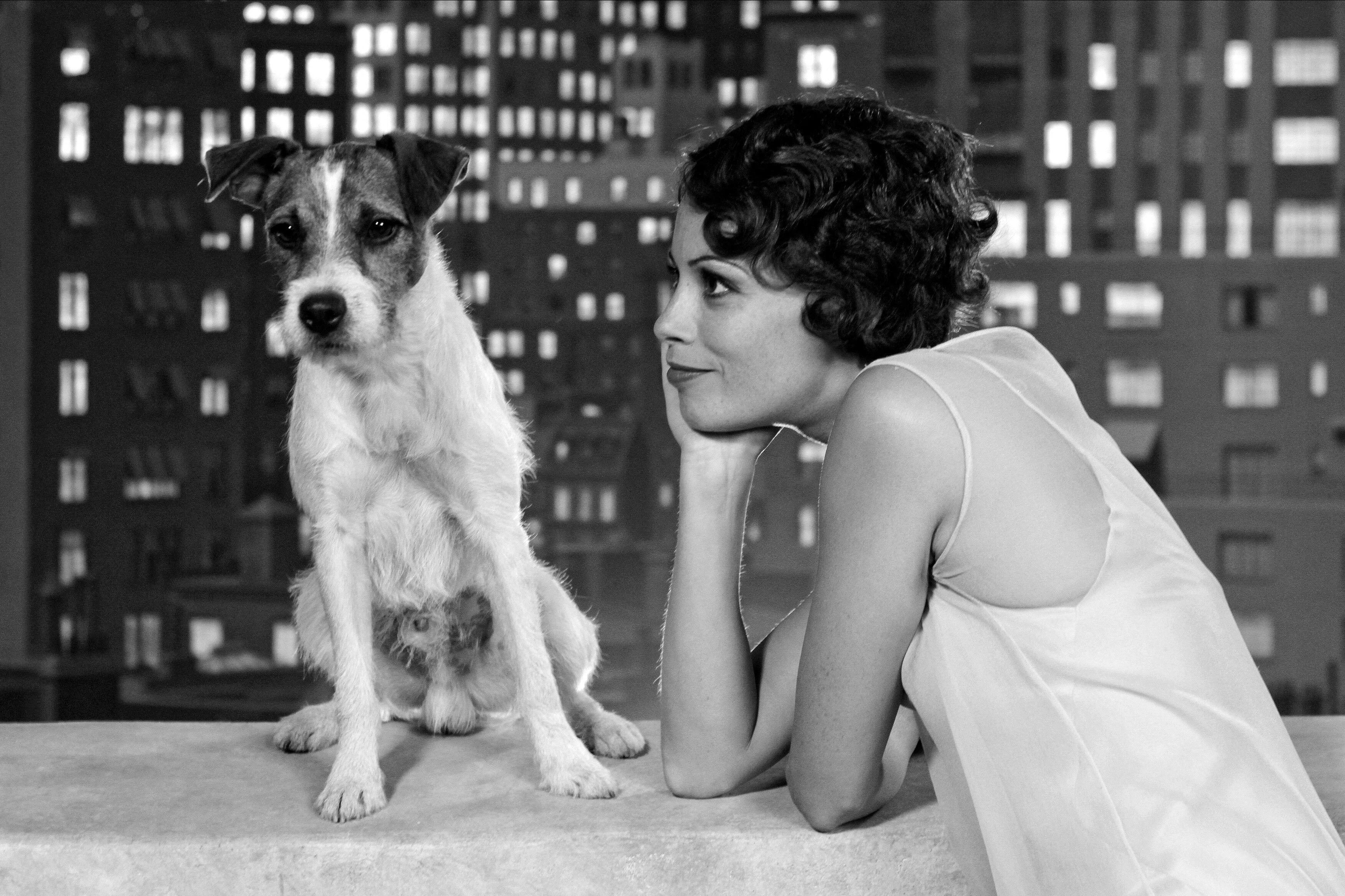 A woman in a 1920s-style dress leans on her hand and smiles at a small terrier sitting beside her, with a blurred nighttime cityscape behind them.