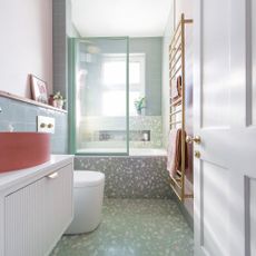 a small bathroom with green wall tiles, a bright coral modern bowl basin and green terrazzo walls and floor around a shower bath