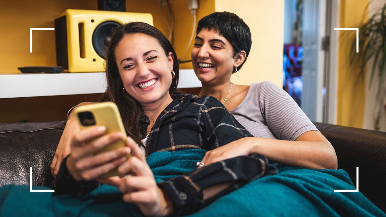 Female couple on the couch, smiling and laughing, holding each other, looking at the best LGBTQ+ podcasts on phone