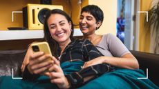 Female couple on the couch, smiling and laughing, holding each other, looking at the best LGBTQ+ podcasts on phone