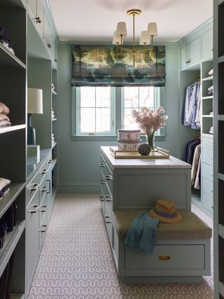 walk in closet painted in aqua blue, blind, central island with drawers, open and closed storage sections, small bench, patterned carpet, pendant light