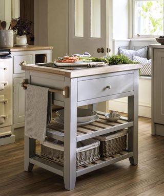 A compact kitchen island with light grey painted wood, a natural oak top, and a single drawer. It includes slatted shelves with wicker baskets for storage and a towel rail on one side. The setup is styled with fresh vegetables, plates, and a window seat in the background for a cosy farmhouse feel.