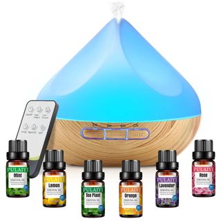 A blue essential oil diffuser with wooden base, remote control, and six small brown bottles of essential oils with black lids in the foreground