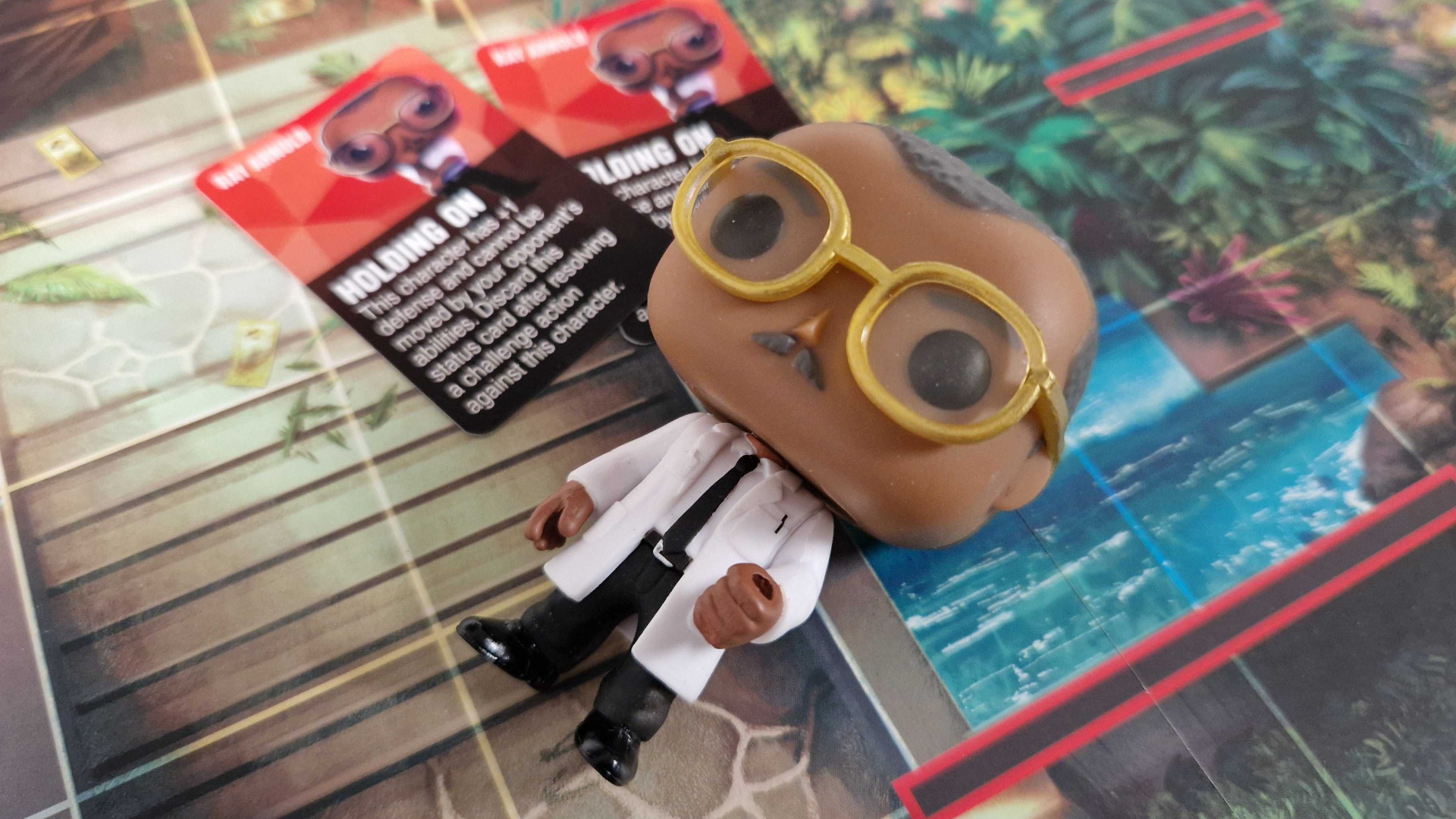 A Ray Arnold Funko figure lying on the Jurassic Park board, with two cards beside it