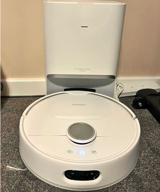 The white round SwitchBot S10 docks at its tall white slim base station on gray carpet against a neutral wall