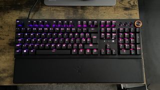 A Razer Huntsman V3 Pro pictured in a slightly dark room, shown on a wooden desk top with purple and pink backlighting behind the keys.