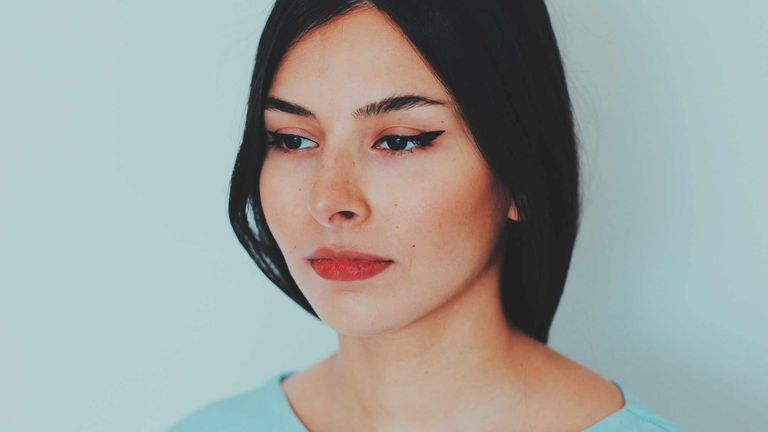 A close up portait of a woman looking away with the perfect eyeliner flicks - has she used these eyeliner hacks? -, you can just see the top of her light blue scoop neck top