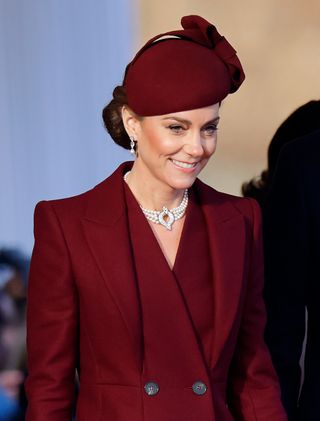 Kate Middleton wearing a maroon coat and pearl choker with a maroon hat and smiling