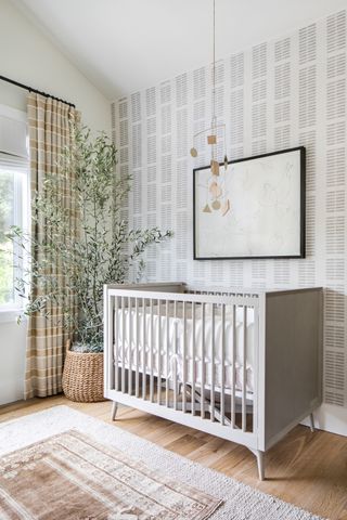 neutral baby girl nursery with grey cot, artwork, graphic wallpaper, rug, tree, neutral check drapes, mobile