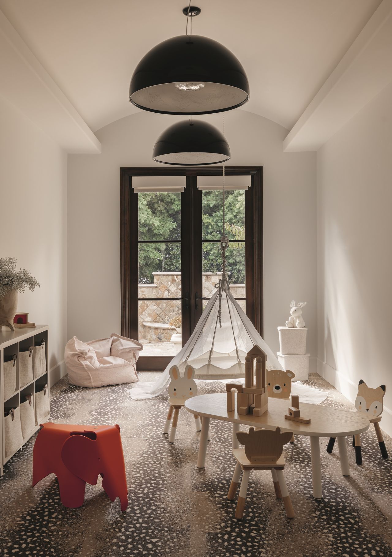 modern playroom with fawn carpet, small table with animal-shaped chairs, orange elephant toy, bean bag, console with storage cubbies, blank pendant lights, and black-framed doors, with tepee in the background