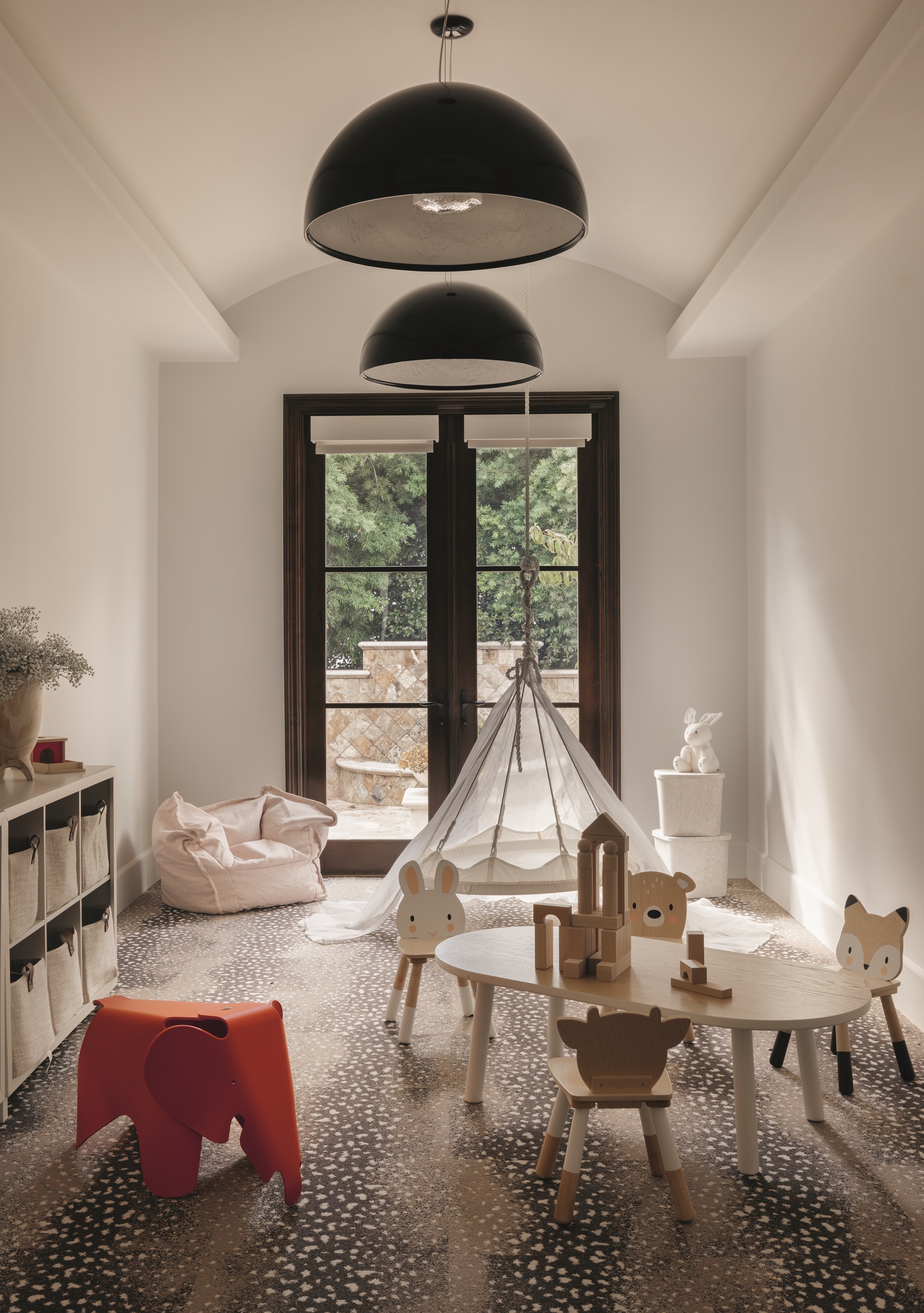 modern playroom with fawn carpet, small table with animal-shaped chairs, orange elephant toy, bean bag, console with storage cubbies, blank pendant lights, and black-framed doors, with tepee in the background