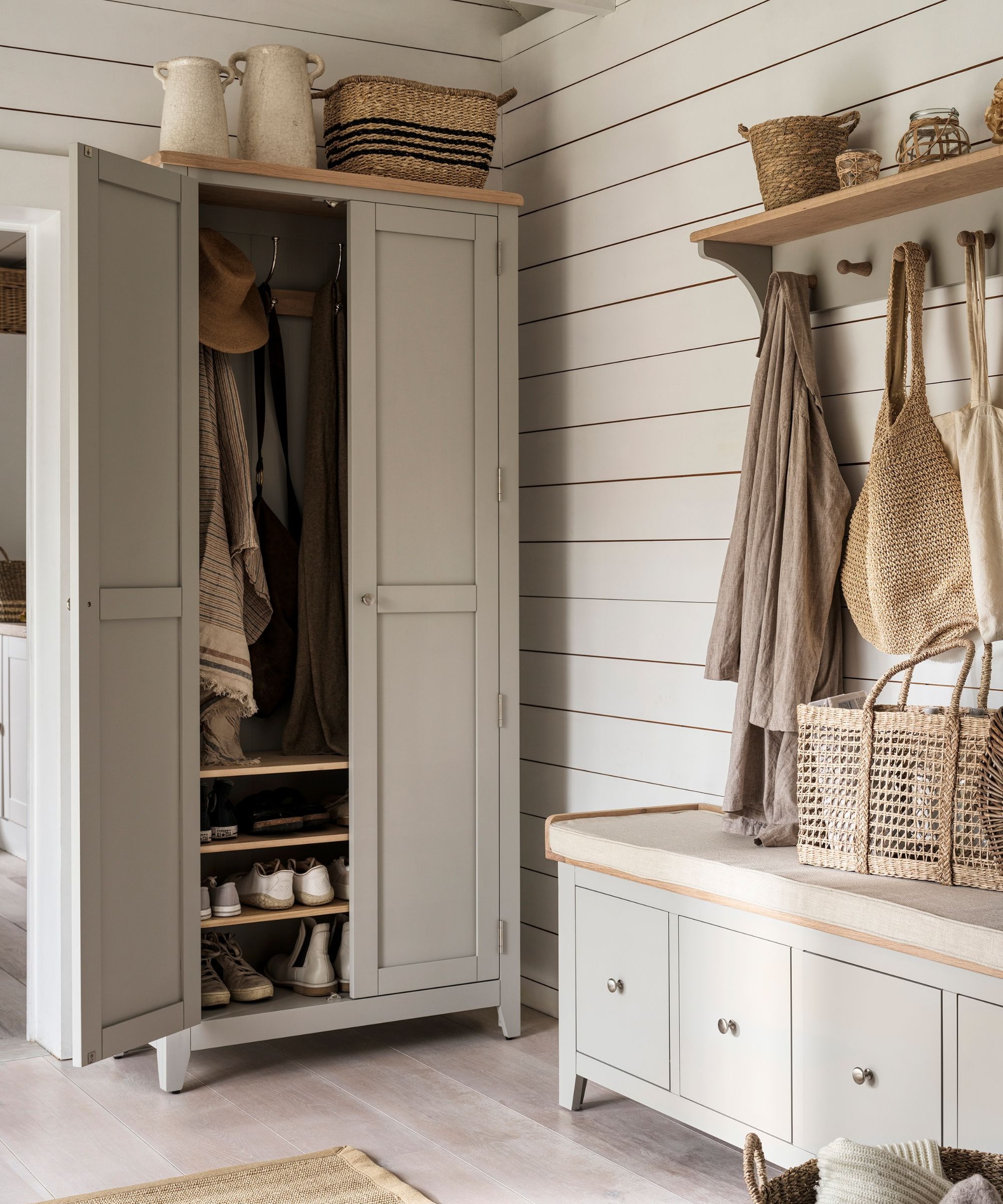 A country-style hallway with a sage green wardrobe open to reveal internal oak shoe shelves and hanging coats, alongside a matching storage bench and wall-mounted coat rack with wooden pegs holding linen jackets and wicker bags on a shiplap-panelled wall.