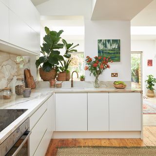 white kitchen with neutral marble worktops, white wall and base units, artwork, wooden floorboards with rug, plants