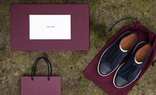 An overview of a pair of black lace up casual shoes with white soles on a burgundy fabric bag next to a burgundy box and shopping bag.