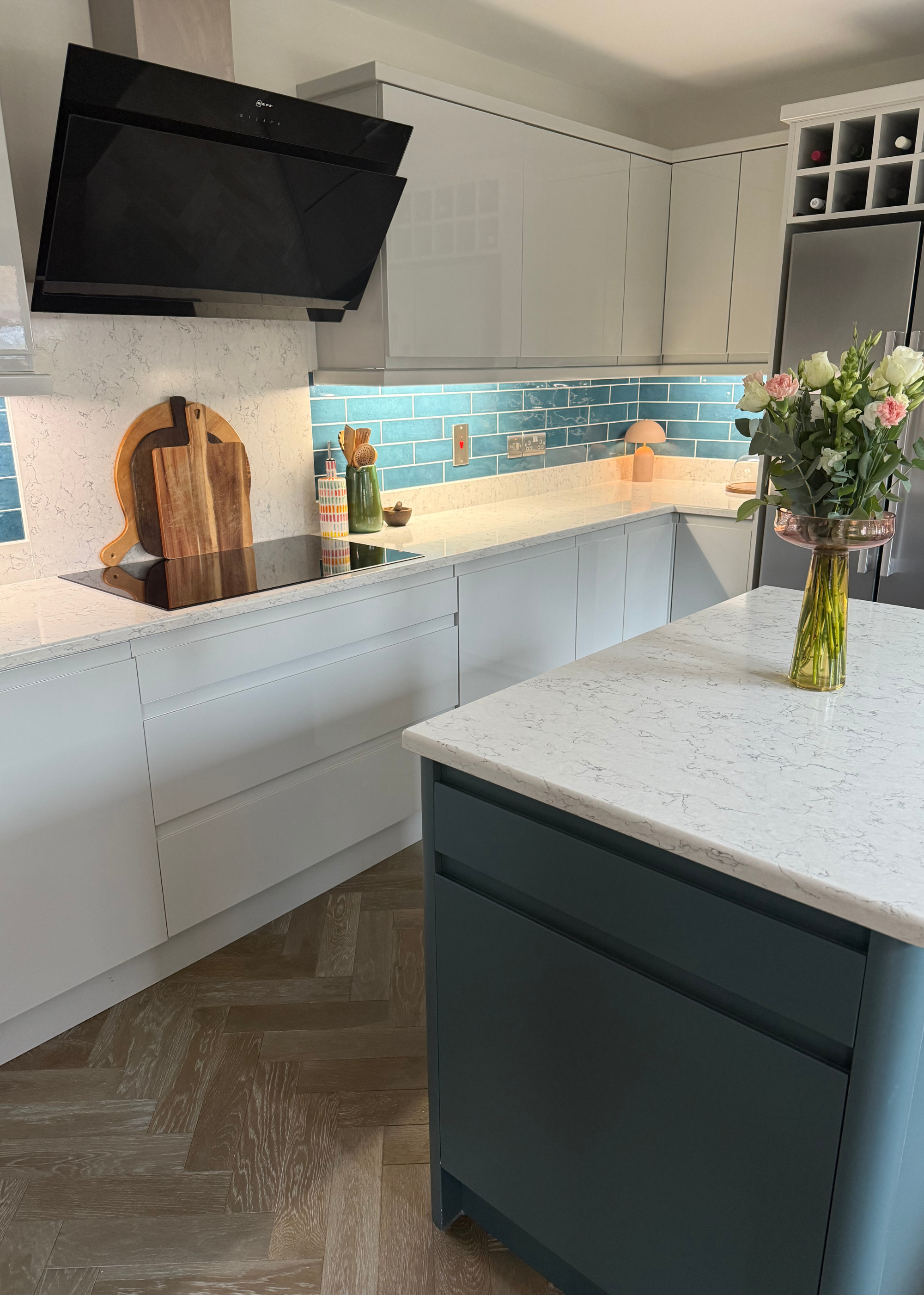 A kitchen with light gray gloss kitchen cabinets with light white and gray-veined quartz countertop with a tiled teal backsplash and matt teal kitchen island with a vase of flowers on top