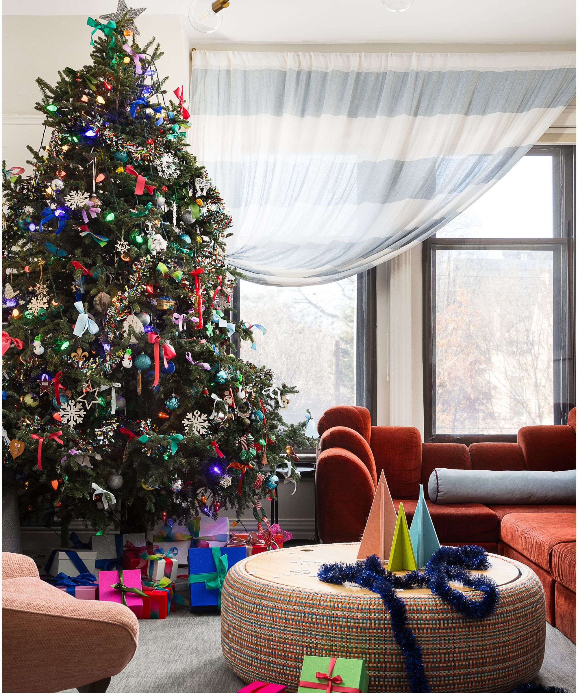 designer Bethany Adams&#039; living room decorated for christmas with a rust velvet couch, ottoman styled with paper trees and a large colorful christmas tree