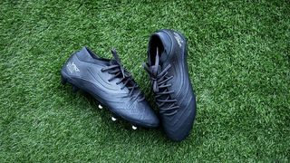 A black pair of football boots, one with white studs showing, lay next to eachother on green artificial grass. Kipsta Viralto IV Pro