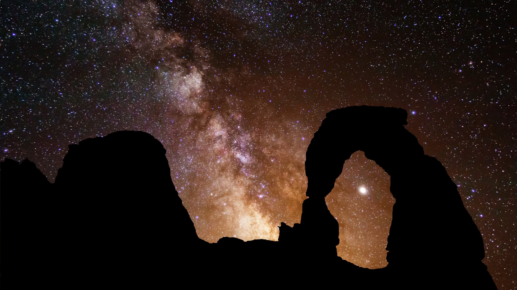 a rock formation in the foreground takes the shape of an arc while the star studded sky behind shows the band of the milky way stretching across the sky.