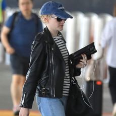 Julia Garner at LAX wearing a blue baseball cap, striped T-shirt, leather jacket, jeans, and Mary Jane flats.