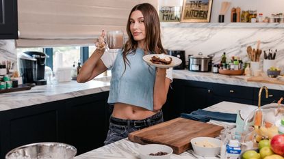 A picture of Hailey Rhode Bieber in her kitchen holding up a coffee cup and plate of pancakes