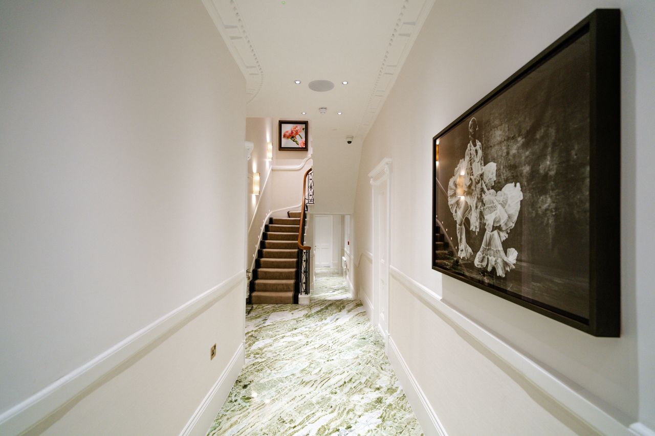 The entrance hallway of a beauty salon is decorated with shiny green and white marble, a large-scale, framed monochromatic portrait of a dancer in an ornate, voluminous dress, and wrought iron and wood details.