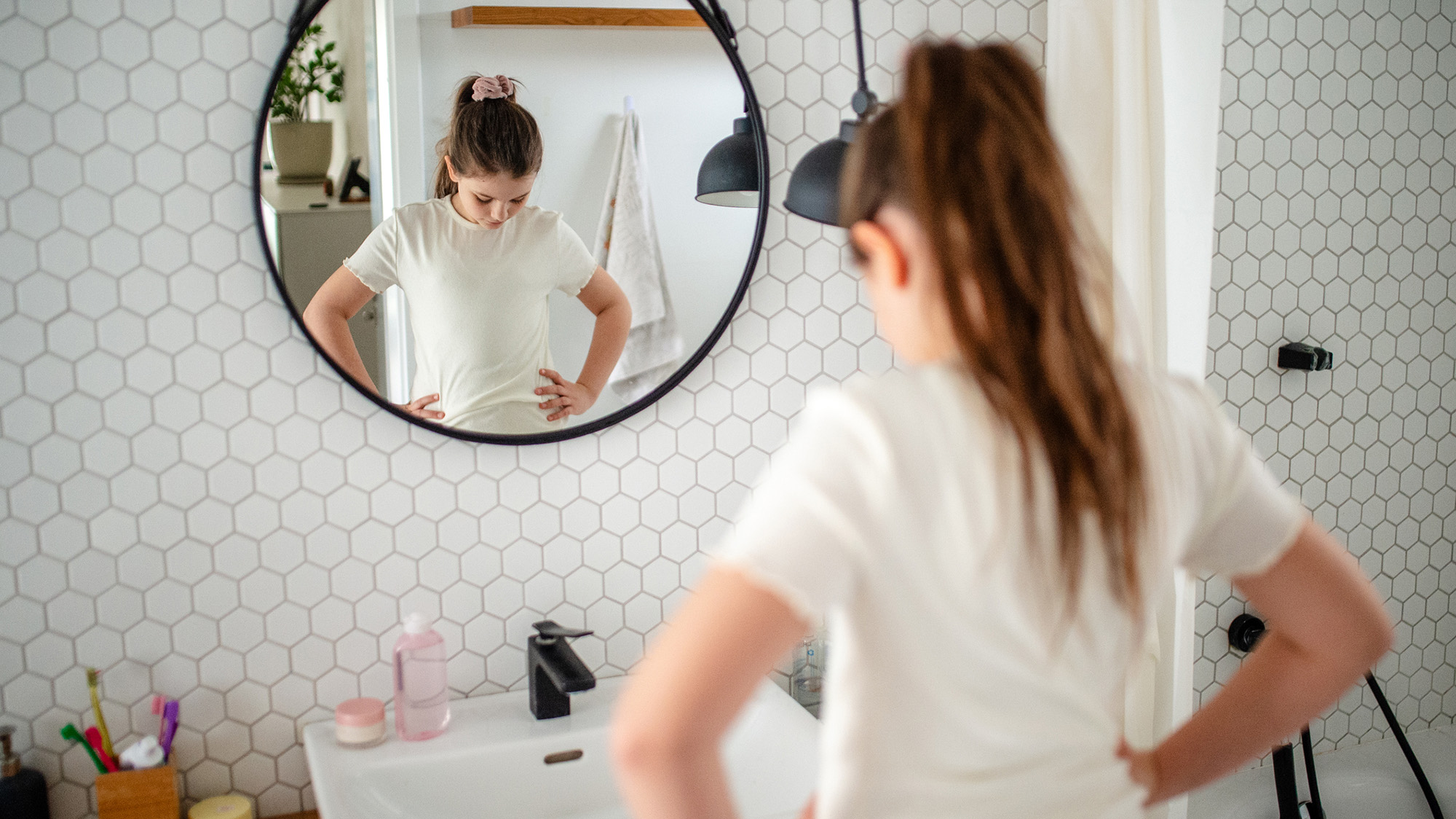 A teen girl with her dark hair in a pony tail stands in front of a mirror.