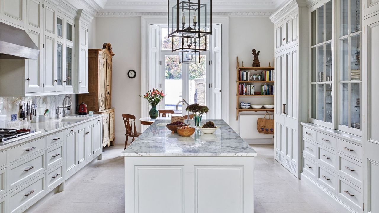A white kitchen with marble island, white and glass cabinets, and wooden shelves and dresser.