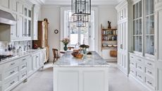 A white kitchen with marble island, white and glass cabinets, and wooden shelves and dresser.