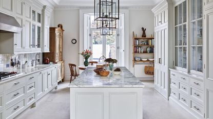 A white kitchen with marble island, white and glass cabinets, and wooden shelves and dresser.