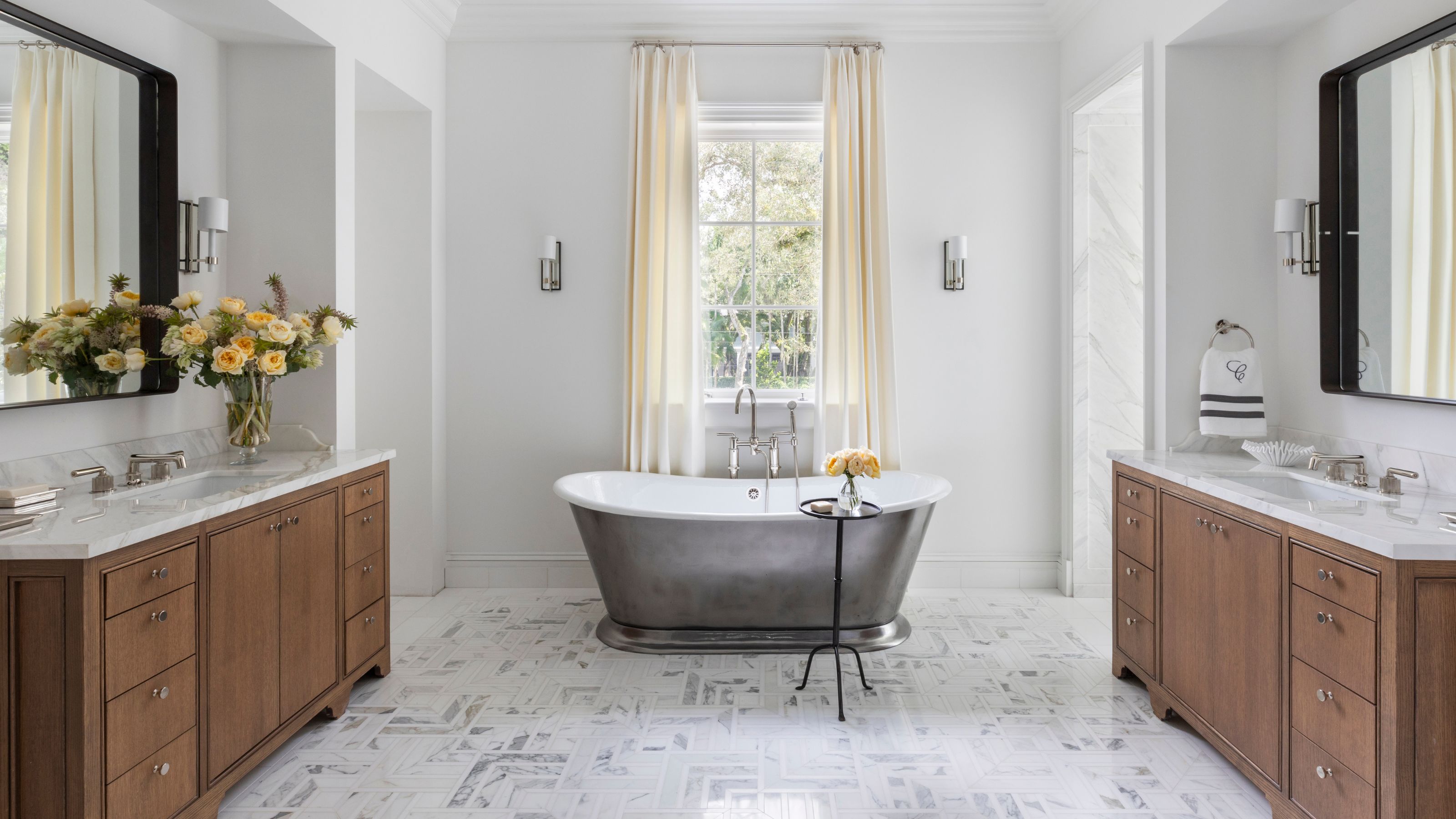 a light white large bathroom with a silver freestanding bathtub positioned under a window with two wooden vanities on either side of the room