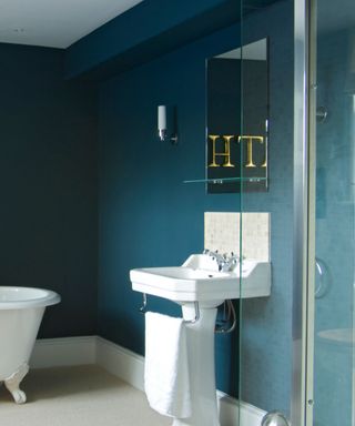 A dark blue bathroom with a glass shower cubicle, white sink with metal towel rail build in, tall wall mounted mirror, and the corner of a freestanding bath with ornate feet