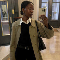 Woman wearing white button-down, black sweater, and car coat, taking a photo of herself in mirror.