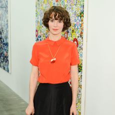 Miranda July wearing bright orange top and black pants in front of colorful canvas