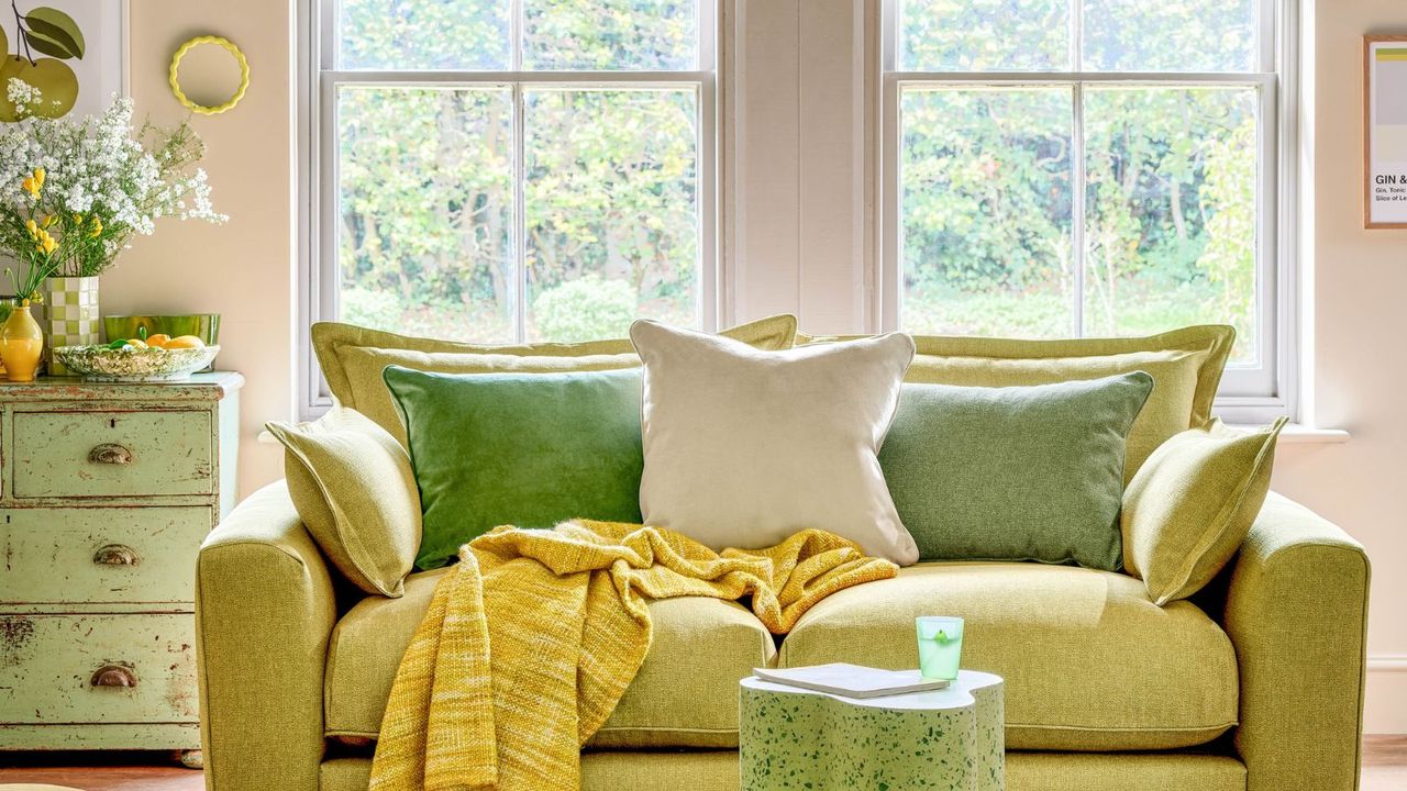 A living room with beige walls with two windows, a distressed green set of drawers with flowers on top, and a green couch in the middle with a yellow throw on it and a green table in front of it.