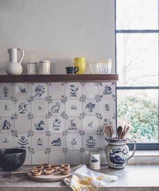 Kitchen worktop with blue and white Delft style wall tiles and crockery, and shelf with glasses and jars.