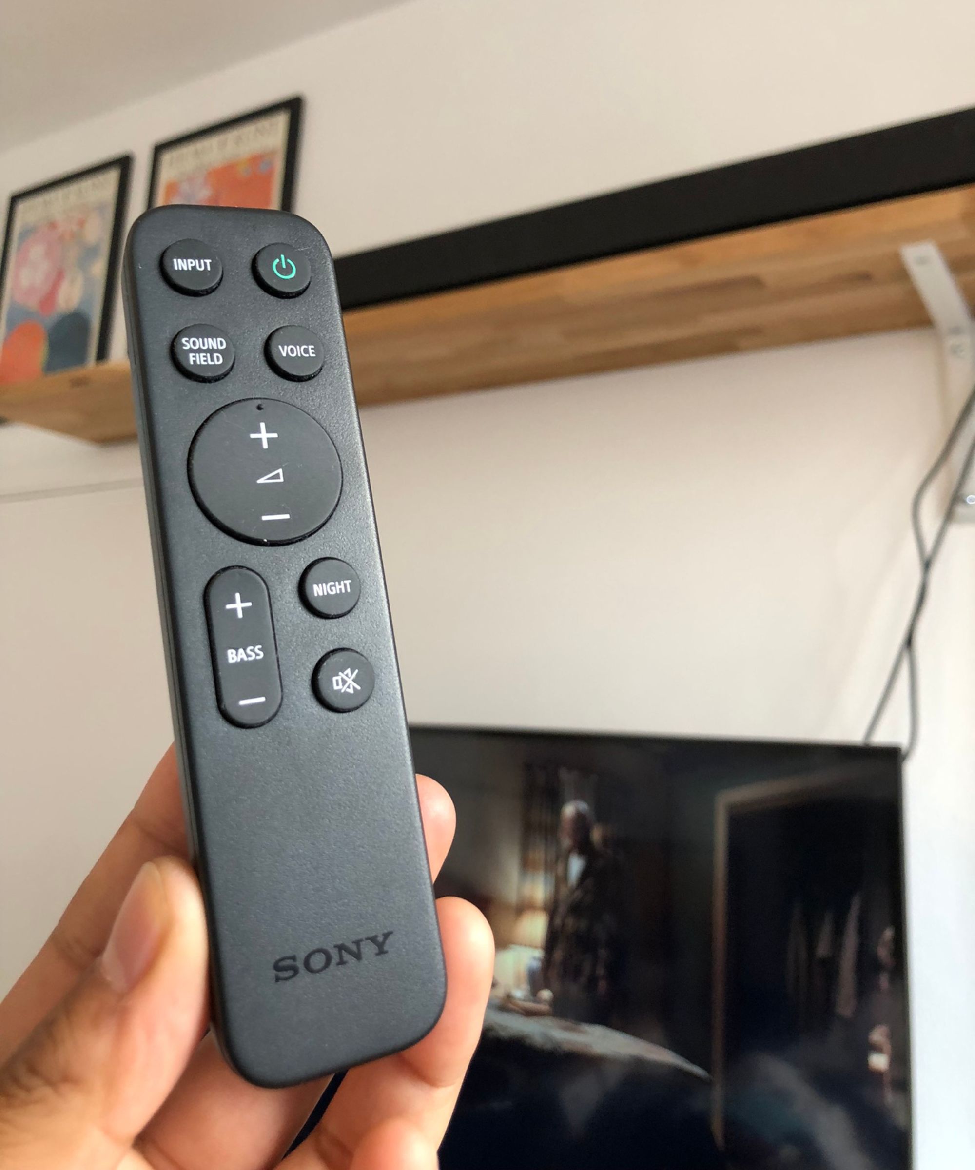 Hand holding the Sony soundbar remote in front of Sony BRAVIA Theater Bar 8 soundbar on wooden shelf, pictured next to art prints and above TV showing Breaking Bad, in Dan Fauzi&#039;s home.