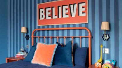 A bedroom with wall covered in a blue striped wallpaper, a sign above the bed reading &#039;believe&#039; and wall lights on either side of the bed