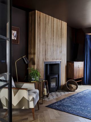 living room with aubergine/brown walls and ceiling, herringbone laid floor, blue rug and drapes, graphic print fabric armchair, wooden slats around log burner, tiled hearth, artwork, retro sideboard, floor lamp, side table