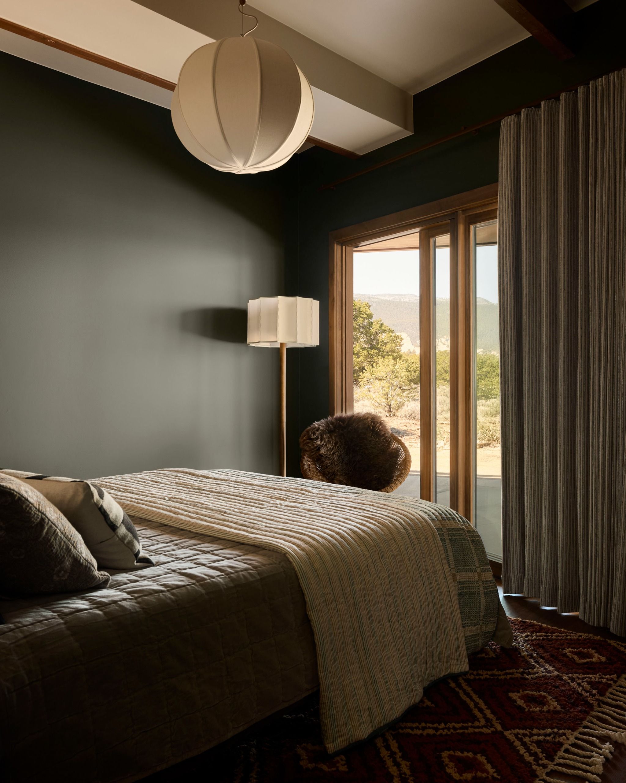 A modern bedroom with deep dark green walls that almost look black, a cream bedspread over the bottom of the made bed with cushions, and a floor lamp in the corner next to the open sliding door